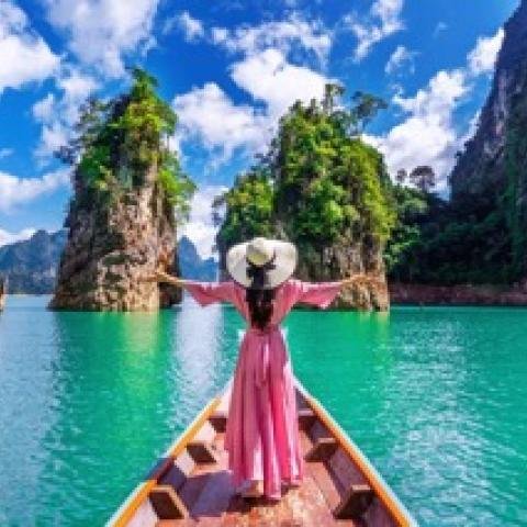 A woman in a pink dress and wide-brimmed hat stands on a boat, arms outstretched. She faces a serene turquoise lake surrounded by lush green cliffs.