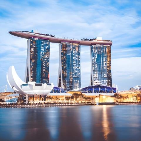 A view of Marina Bay Sands in Singapore at dusk, reflecting in the calm water. The sky is a soft blue, accentuating the futuristic cityscape.