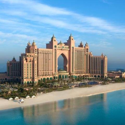 A luxurious oceanfront resort with expansive sandy beaches, lush greenery, and grand architecture featuring an arched center, under a clear blue sky.