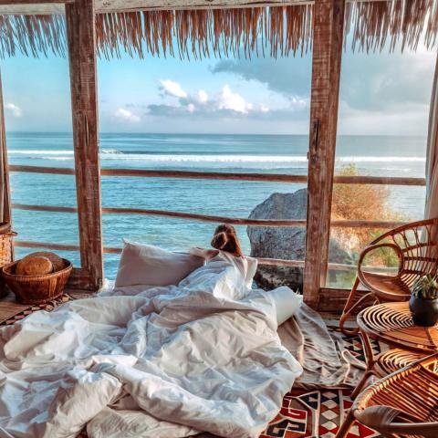 Cozy bedroom with a bed facing large windows overlooking the ocean. Wooden furniture and wicker chairs create a rustic, serene ambiance.