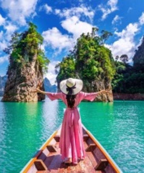 A woman in a pink dress and wide-brimmed hat stands on a boat, arms outstretched. She faces a serene turquoise lake surrounded by lush green cliffs.