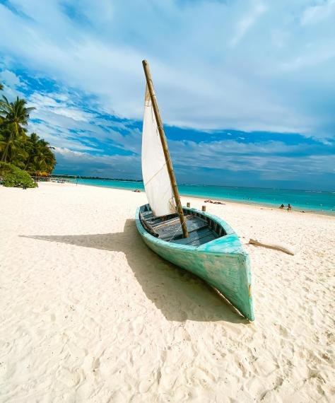 A light blue boat with a white sail rests on a sunlit beach with turquoise waves in the background. Palm trees line the shore under a partly cloudy sky.