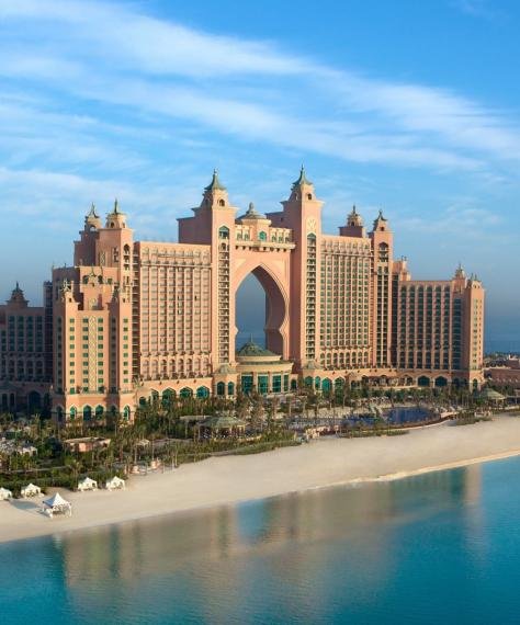 A luxurious oceanfront resort with expansive sandy beaches, lush greenery, and grand architecture featuring an arched center, under a clear blue sky.