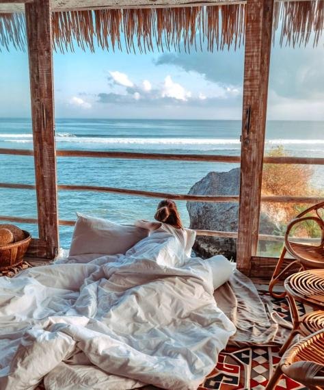 Cozy bedroom with a bed facing large windows overlooking the ocean. Wooden furniture and wicker chairs create a rustic, serene ambiance.