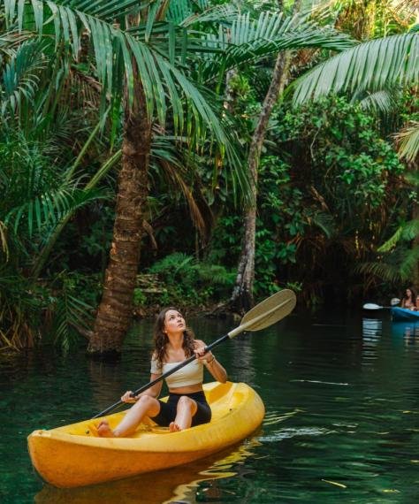 A woman in a yellow kayak paddles through lush, tropical greenery, exuding a sense of adventure. Behind her, another kayak can be seen on the water.