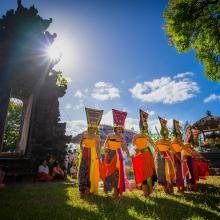 Traditional Balinese dancers in vibrant costumes perform in sunlight, flanked by temple gates and onlookers. The scene feels lively and celebratory.