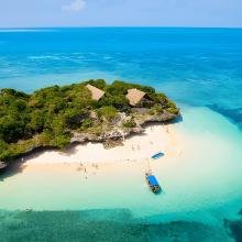 A small, lush green island with thatched-roof huts sits in vibrant turquoise waters. A sandy beach curves around, with a boat anchored near the shore.