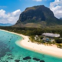 Aerial view of a tropical beach with turquoise waters and a lush coastline. A towering mountain rises in the background under a partly cloudy sky.