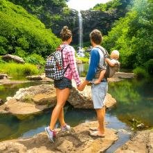 A couple with backpacks stands on rocks by a stream, holding a baby, gazing at a waterfall surrounded by lush greenery, conveying adventure and tranquility.