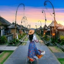 A woman in a flowing dress and hat walks down a charming Balinese village path at sunset, surrounded by traditional houses and festive decorations.