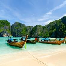 Long-tail boats float on turquoise water, moored near a sandy beach. Lush green cliffs rise in the background under a clear blue sky, conveying tranquility.