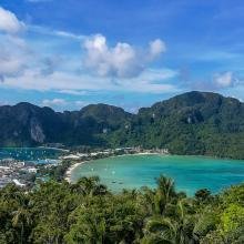 Scenic view of a tropical bay with clear turquoise water, surrounded by lush green hills and a vibrant town, under a bright blue sky with fluffy clouds.
