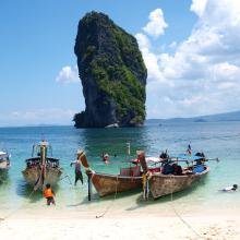Tropical beach scene with colorful boats docked on clear turquoise water, people swimming, and a lush rock formation under a bright blue sky.