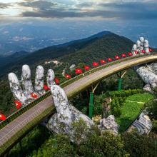 A stunning curved bridge, supported by giant stone hands, spans a lush green mountainside. Red flags line the bridge, creating a dramatic, majestic scene.