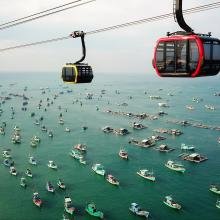 Aerial view of two brightly colored cable cars gliding over a vast blue ocean filled with numerous fishing boats, creating a lively and bustling scene.