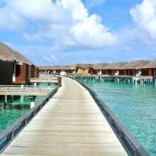 Wooden pathway leads to thatched-roof overwater bungalows on serene turquoise waters under a vibrant blue sky with fluffy clouds. Peaceful and tropical.
