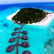 Aerial view of a tropical island with lush greenery, white sandy beaches, and turquoise water. Overwater bungalows extend into the ocean, conveying tranquility.