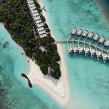 Aerial view of a tropical island resort with lush greenery and white sandy beaches. Overwater bungalows on a turquoise sea resemble a palm frond.