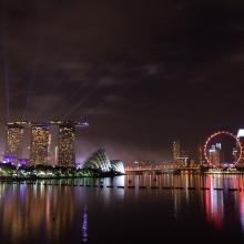 Night cityscape of Singapore featuring Marina Bay Sands, an illuminated Ferris wheel, and glowing skyline. Reflections shimmer on calm water, creating a vibrant, dramatic scene.