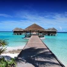 A wooden pier leads to a thatched-roof structure over turquoise waters, under a deep blue sky. Palm leaves and sand border the scene, evoking tranquility.