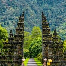 Tall, ancient Balinese gate flanked by lush green trees and mountains. The pathway leads through the open stone structure, conveying tranquility and grandeur.