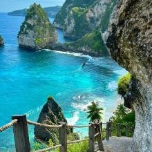 A scenic view of a rocky coastal landscape with turquoise water, cliffs, and lush greenery. A wooden railing lines a stone path descending to the beach.