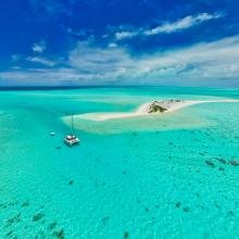 A tranquil scene of a catamaran floating on crystal-clear turquoise waters near a small sandy island under a vibrant blue sky with scattered clouds.
