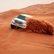 A white SUV powers through a desert, kicking up a dramatic wave of orange sand. The expansive dunes create a thrilling, adventurous scene.