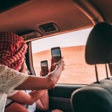 Two people in a car, one wearing a red and white headscarf, take photos with phones through the window, capturing a desert landscape.