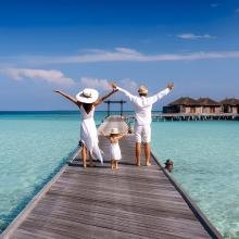 Family in white outfits and hats joyfully stretches arms on a wooden pier over turquoise ocean, facing overwater bungalows under a clear, sunny sky.