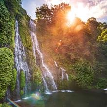 Sunlit waterfall cascades down lush, green cliffs surrounded by dense forest, with sunlight creating a warm, serene atmosphere and lens flares.