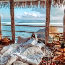 A serene room with a wooden interior features a person in white bed linens looking out at a clear ocean view. Wicker chairs and a colorful rug add warmth.