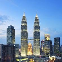 Twin skyscrapers at dusk with a glowing sky bridge, surrounded by city buildings. Sky is a deepening blue, conveying a serene evening ambiance.
