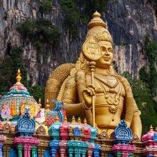 Golden statue of a deity stands majestically against a rocky, forested backdrop. In front, a colorful, intricately decorated temple roof is visible.