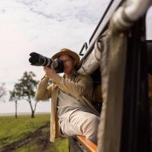 Man in safari hat uses a large camera lens from a vehicle, capturing nature. Open savannah and trees in the background under a cloudy sky. Adventurous tone.