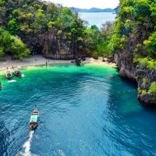 A small boat navigates a vibrant turquoise bay, surrounded by lush, towering cliffs. A sandy beach is nestled between the cliffs, with several people enjoying the serene landscape.