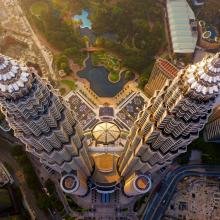 Aerial view of two towering skyscrapers with intricate spires, surrounded by greenery and urban landscape. The scene is bathed in warm, golden sunlight.