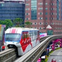Urban landscape with a red and white monorail traveling on an elevated track amidst tall buildings and lush trees; conveys a sense of modernity and efficiency.