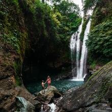 A vibrant waterfall cascades into a green pool surrounded by lush foliage. Two people stand on a rock in the foreground, exuding adventure and serenity.