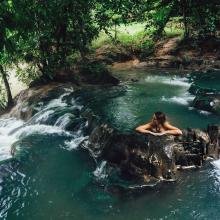 A person relaxes in a natural pool surrounded by lush greenery and cascading waterfalls. The scene is tranquil and idyllic, evoking a sense of serenity.