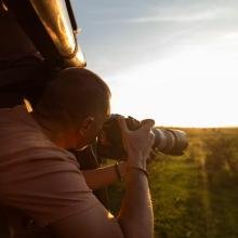 A photographer leans out of a vehicle window, aiming a large camera at a sunset over a grassy field. The scene conveys focus and serenity.