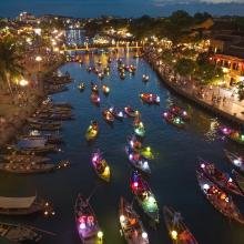 A night view of a river, crowded with colorful lantern-lit boats. Bright lights and lush greenery line both banks, creating a festive atmosphere.